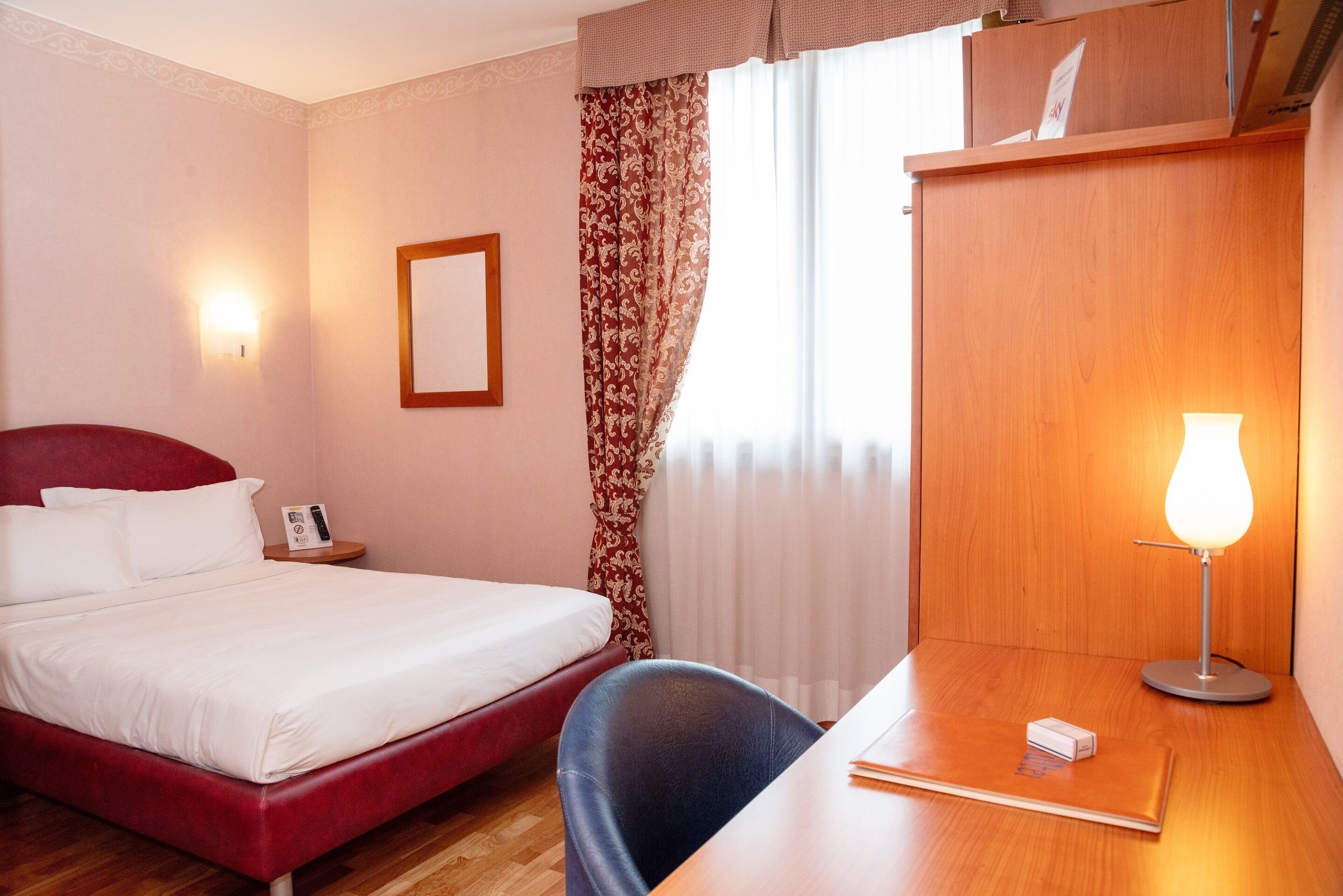 Habitación de hotel con una cama individual, un escritorio con lámpara, una silla, una mesita de noche y una ventana con cortinas.
