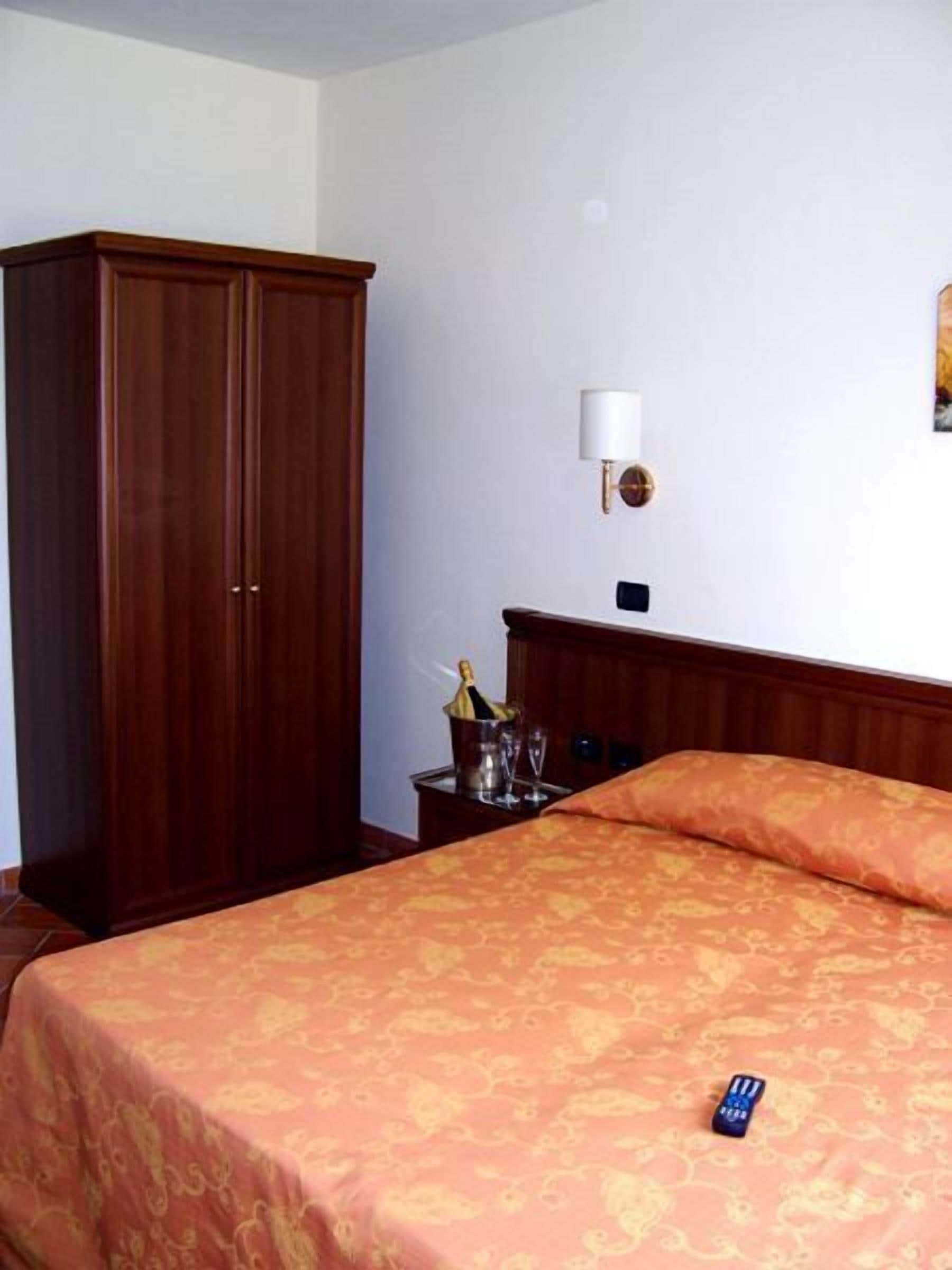 Ein Hotelzimmer mit großem Holzschrank, einem Nachttisch mit Flasche und Gläsern sowie einem Bett mit gemustertem Überwurf.