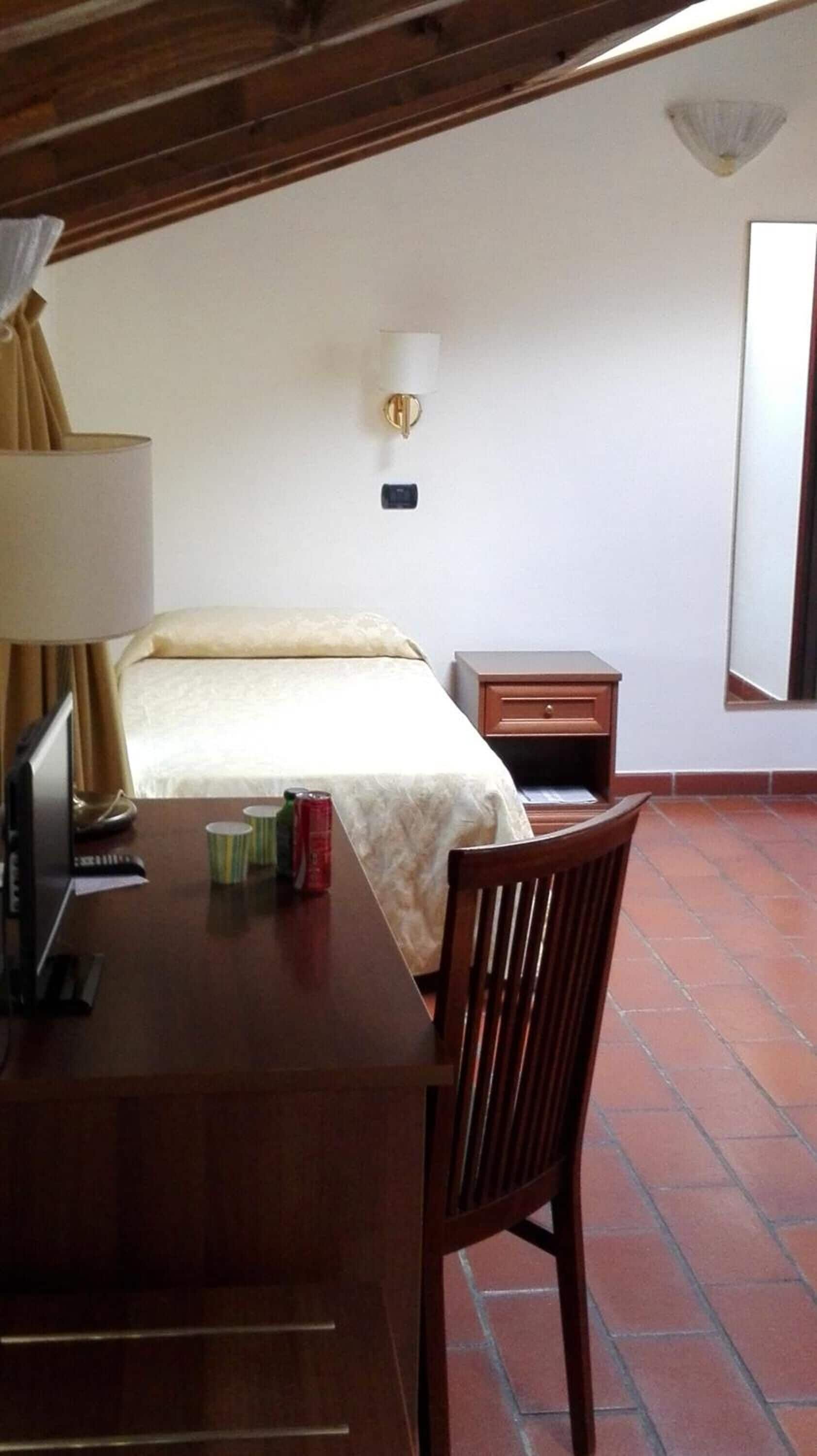 Habitación de hotel con una cama, un escritorio con lámpara, una silla, una mesita de noche y un aplique de pared.