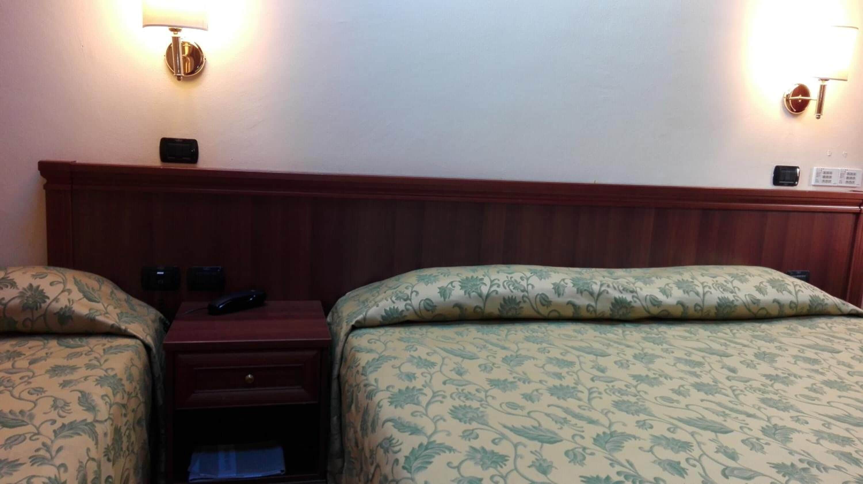 Habitación de hotel con cabecera de madera, dos camas individuales con ropa de cama floral, mesitas de noche con lámparas y una pequeña cómoda.