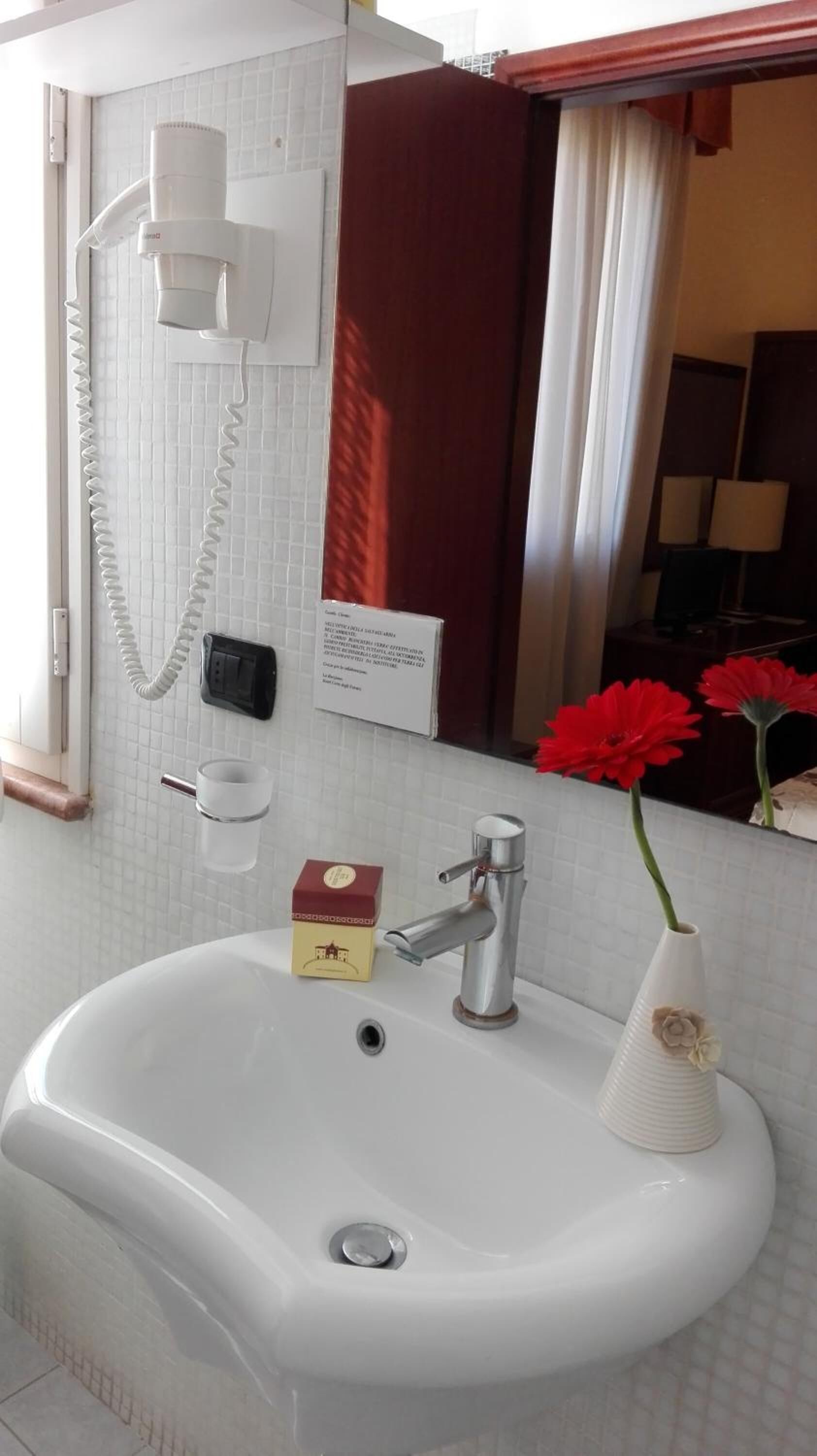 Ein Badezimmer mit einem weißen Waschbecken, einem Spiegel, einem Handtuchhalter, einem Telefon und einer roten Blume in einer Vase.