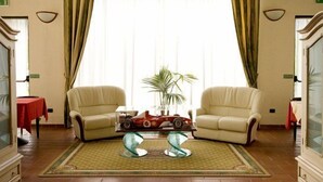 A living room with two beige armchairs, a glass coffee table with a red toy car, a green patterned rug, and a large window with curtains. - Corte degli Estensi (Formigine)