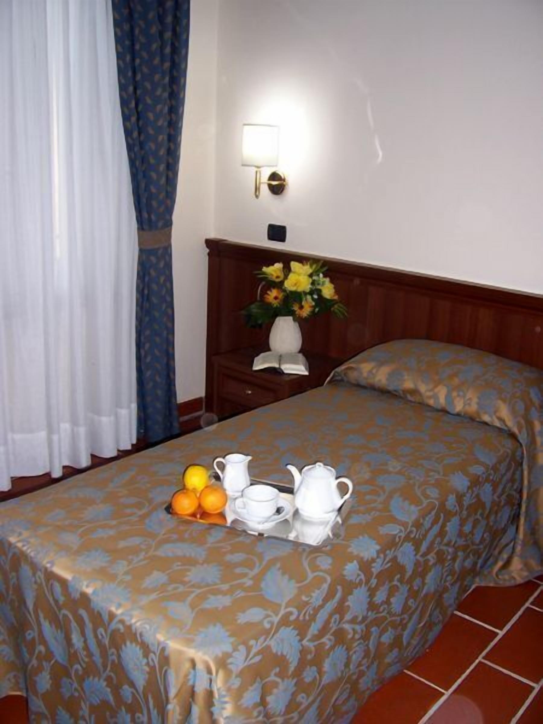 A neatly made bed with a floral bedspread, a bedside table with a lamp, a vase with flowers, and a tray with fruits and tea set on the bed.