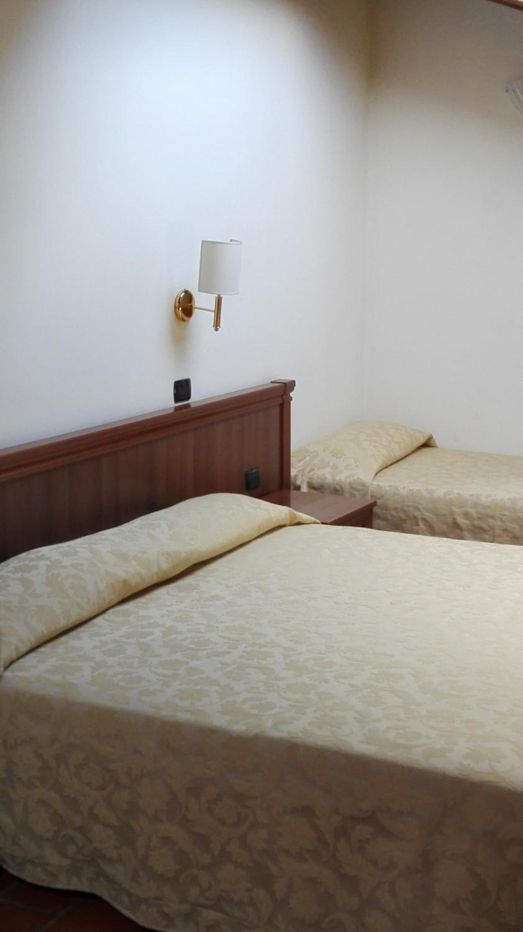 Quarto de hotel com duas camas, cabeceira de madeira, mesa de cabeceira e luminária na parede.