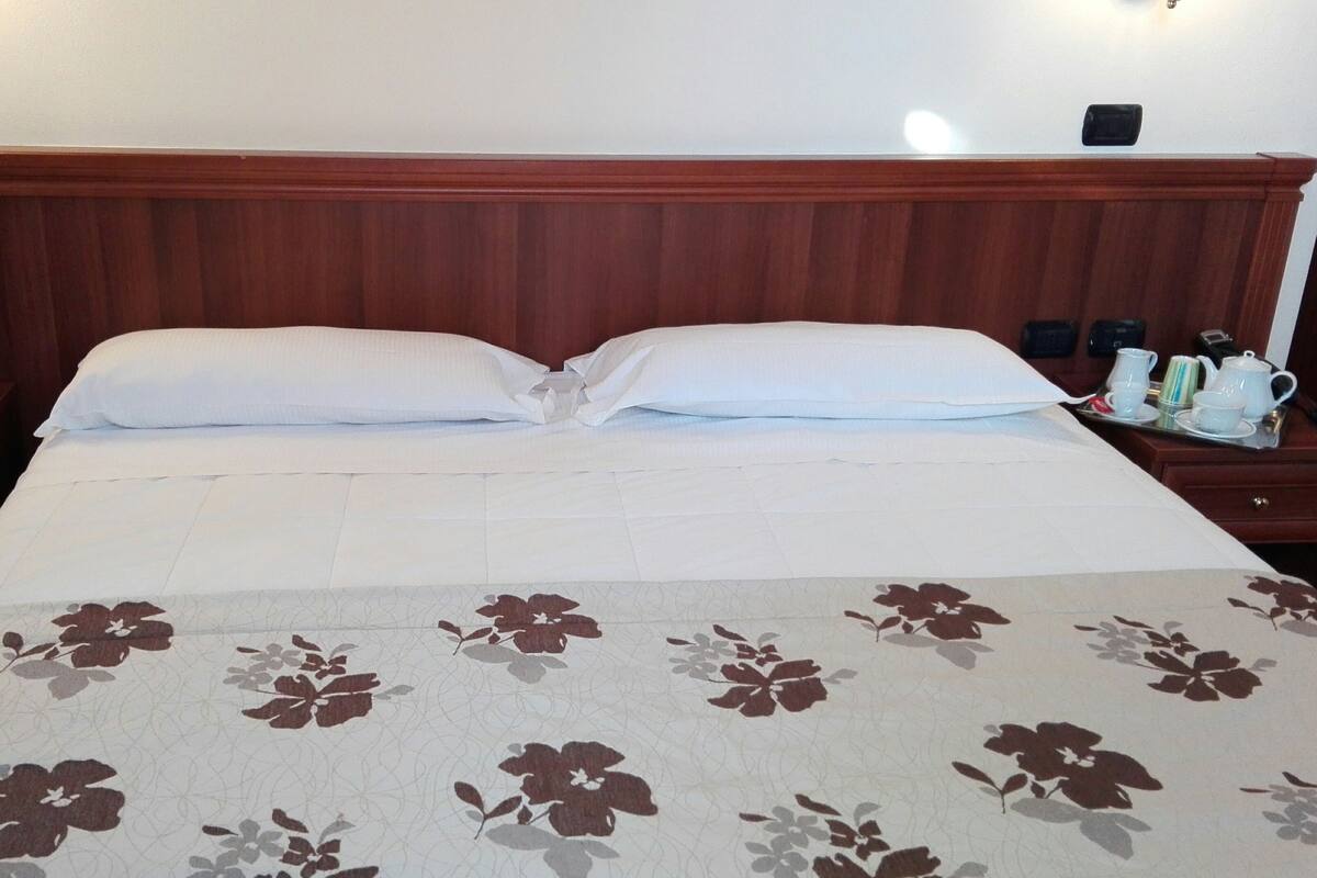 Hotellihuone, jossa on suuri sänky, puinen pääty, kaksi yöpöytää lamppuineen, maljakko kukkineen ja tarjotinalus teekupit ja -lautaset.