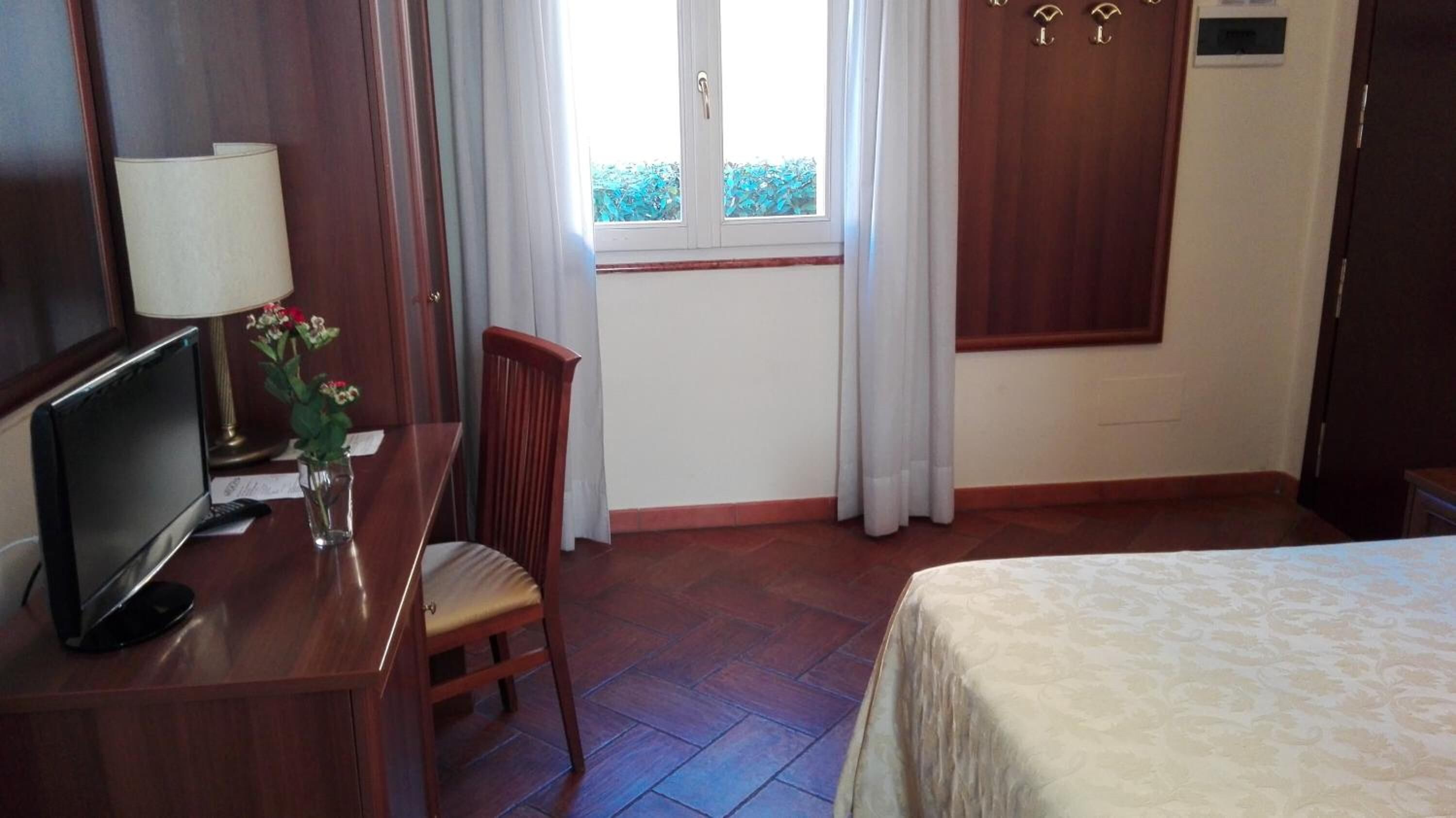 A hotel room with a bed, a desk with a TV, a chair, a window with curtains, and a door.