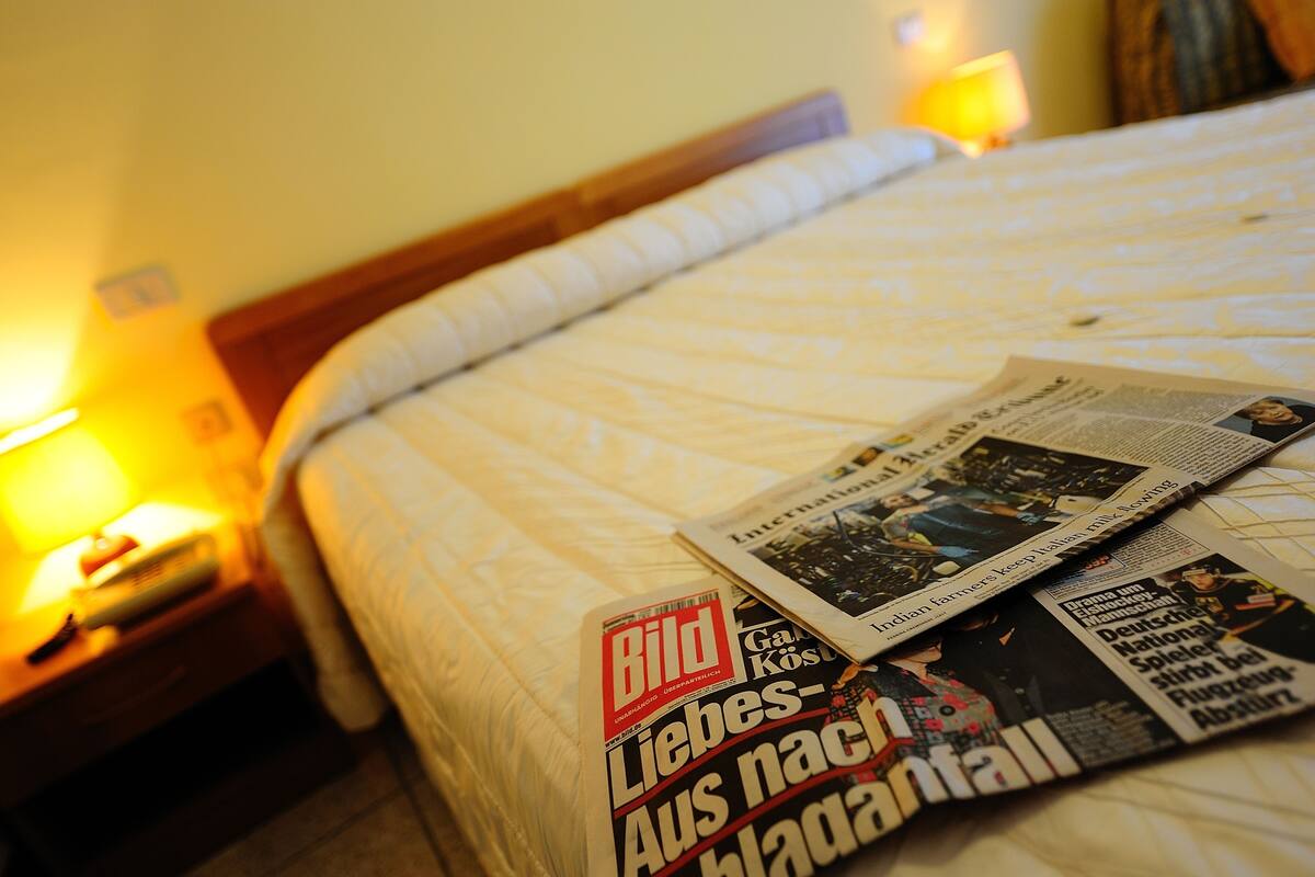 Una cama con periódicos y una lámpara sobre una mesita de noche.