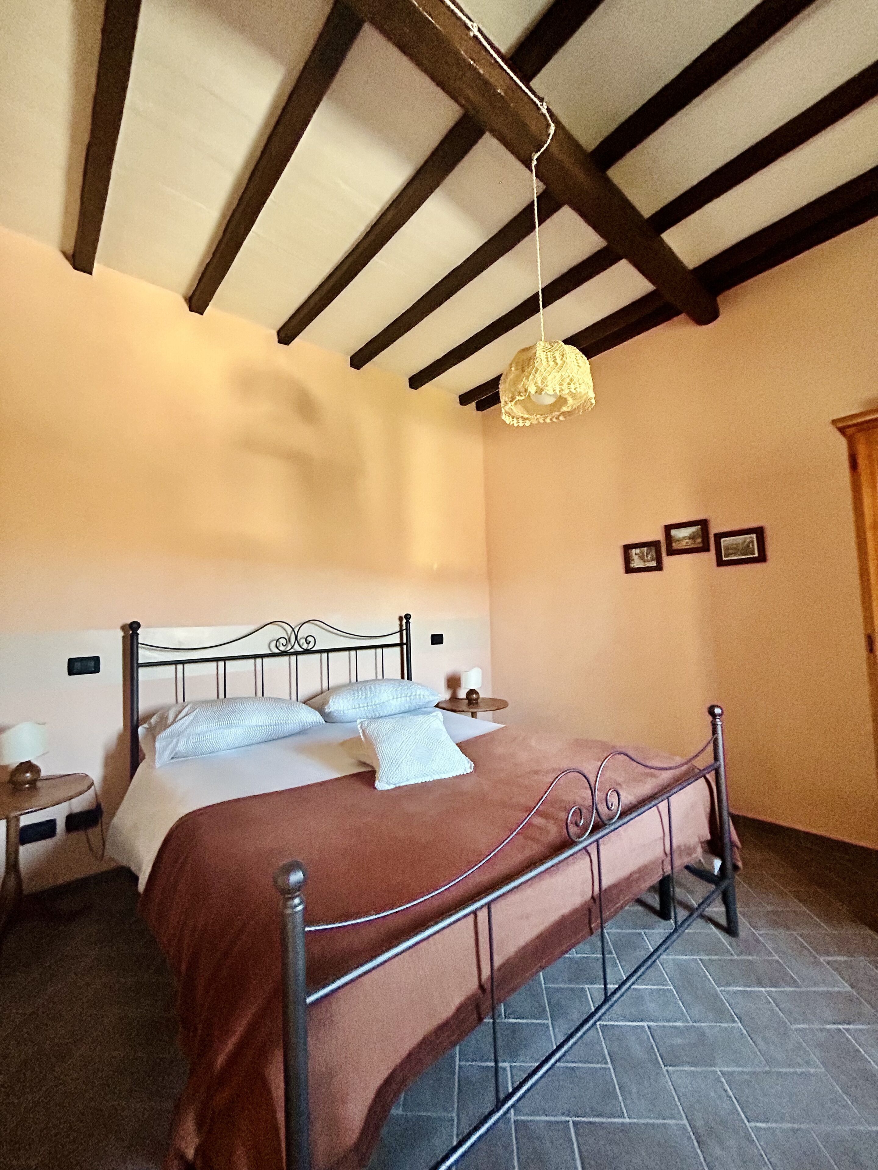 A bedroom with a metal bed frame, a brown bedspread, a hanging light fixture, a bedside table with a lamp, and a small round table with a candle.