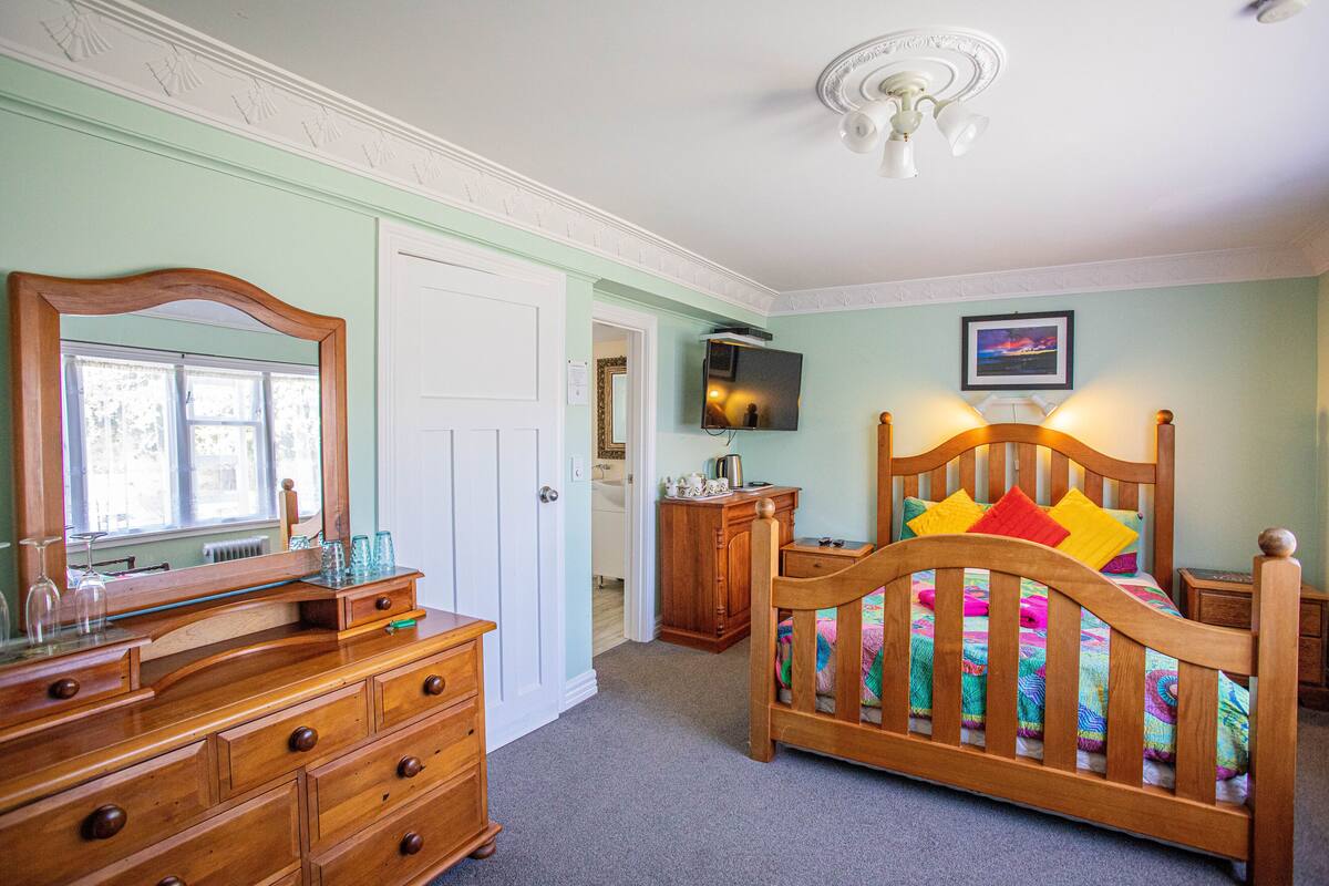 Une chambre à coucher avec un lit en bois, une commode, un miroir, un téléviseur et une porte donnant sur une autre pièce.