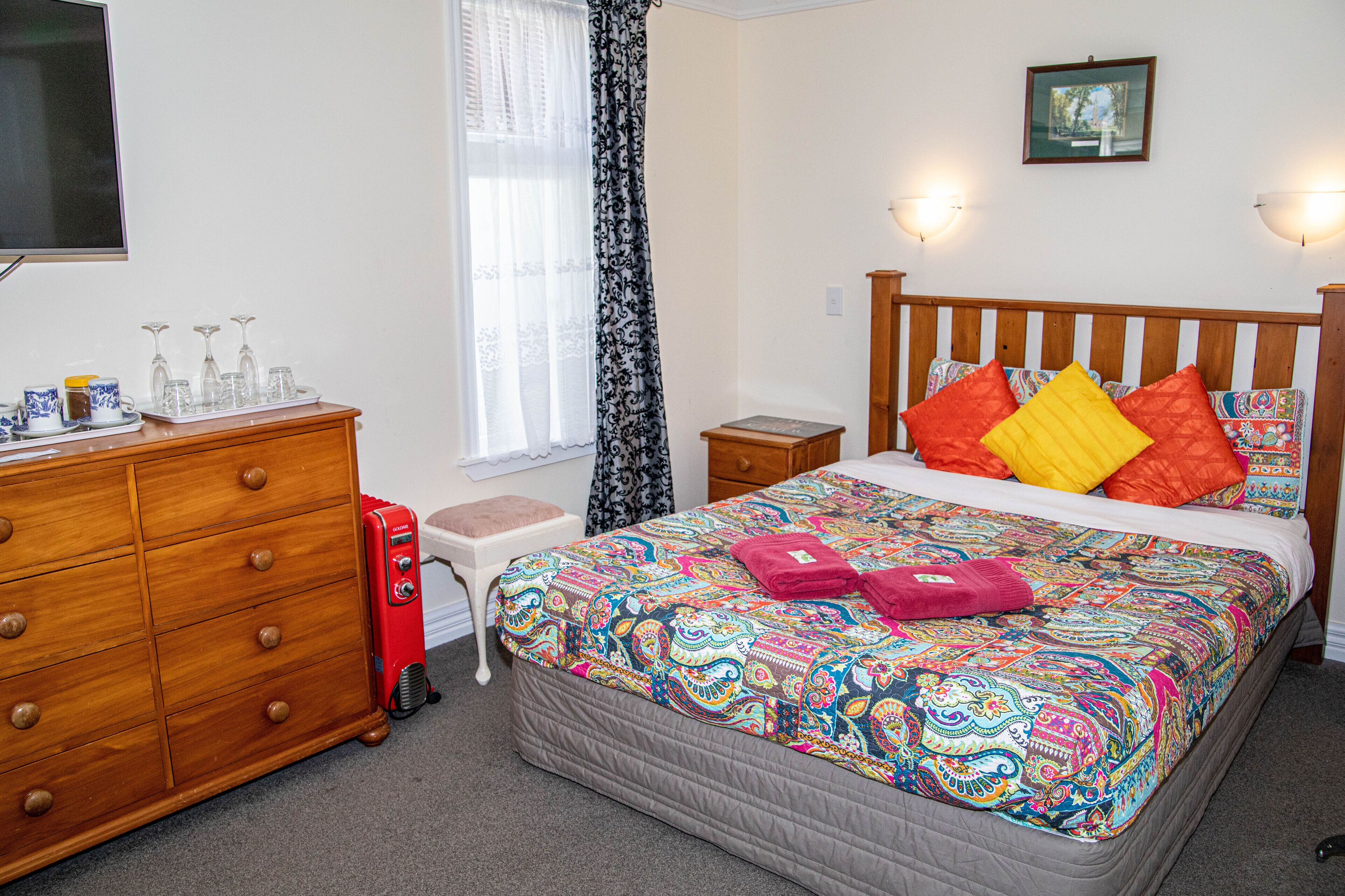 Un dormitorio con cómoda de madera, mesita de noche, cama con ropa de cama colorida, ventana con cortinas y televisor montado en la pared.