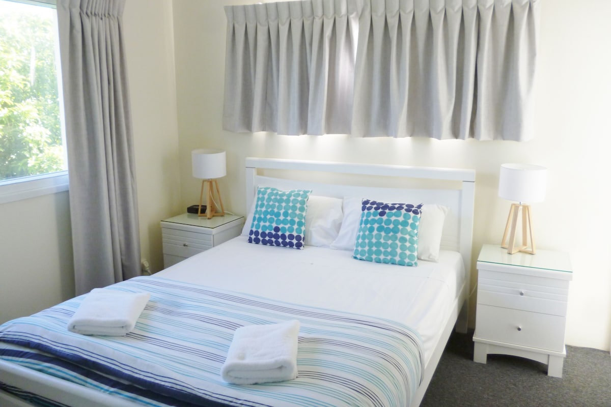 Een hotelkamer met een bed, twee nachtkastjes, een raam met gordijnen en uitzicht op groen.