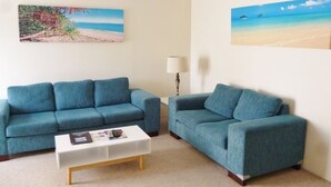 A living room with two teal sofas, a white coffee table, and two beach-themed paintings on the walls. - Le Lavandou Holiday Apartments (Broadbeach)