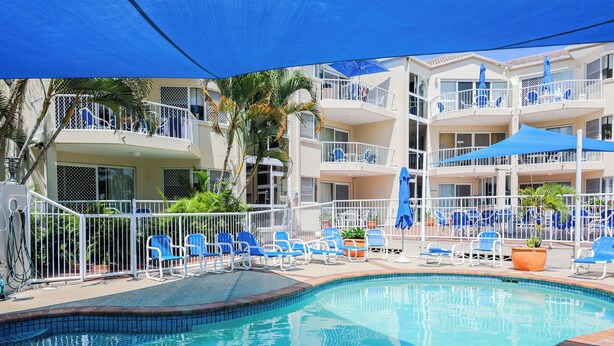 A swimming pool with blue chairs and umbrellas, surrounded by a white fence and a multi-story building with balconies. - Le Lavandou Holiday Apartments (Broadbeach)