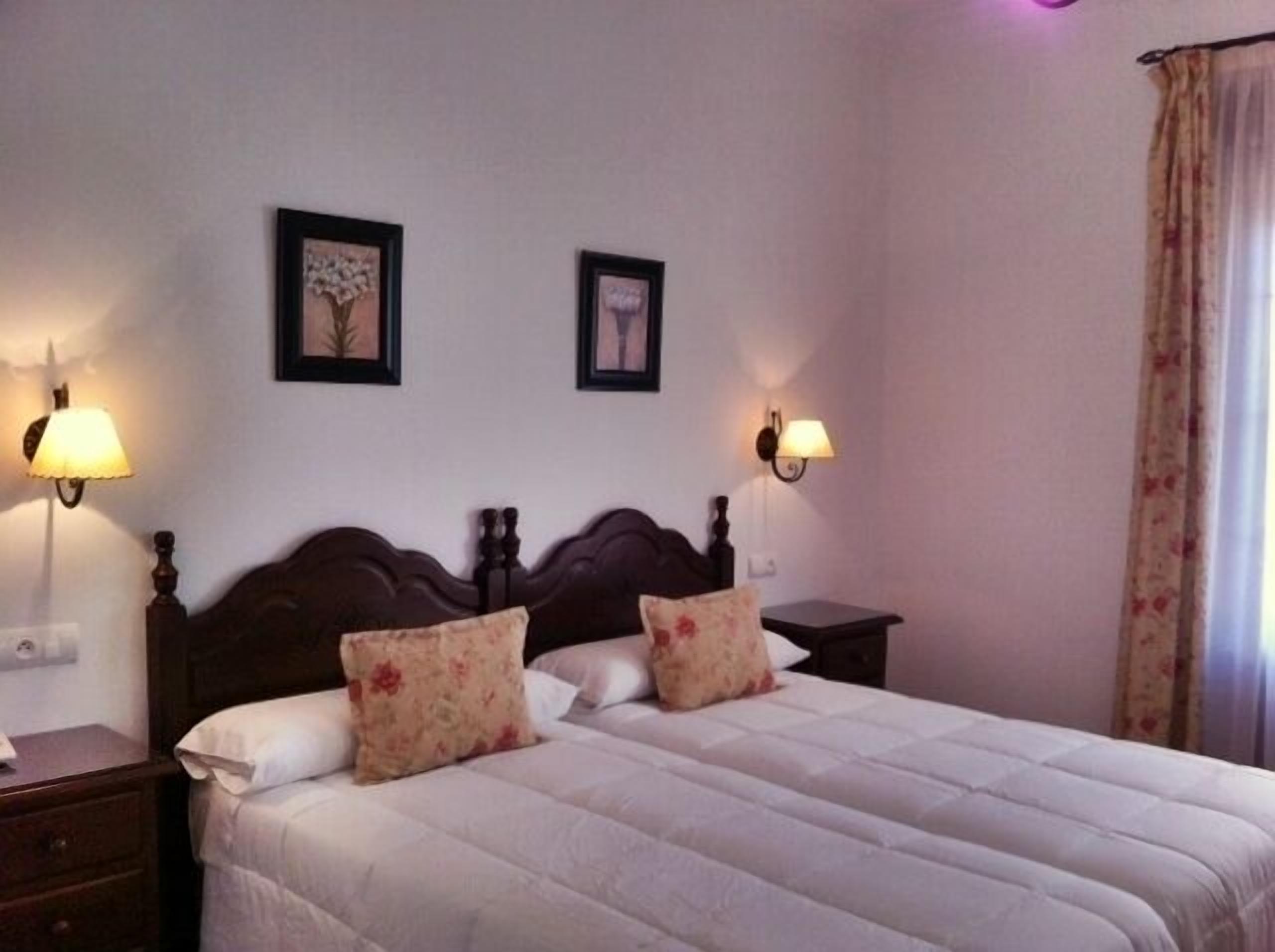 A double bed with floral pillows, two wall-mounted lamps, and two framed pictures on the wall.