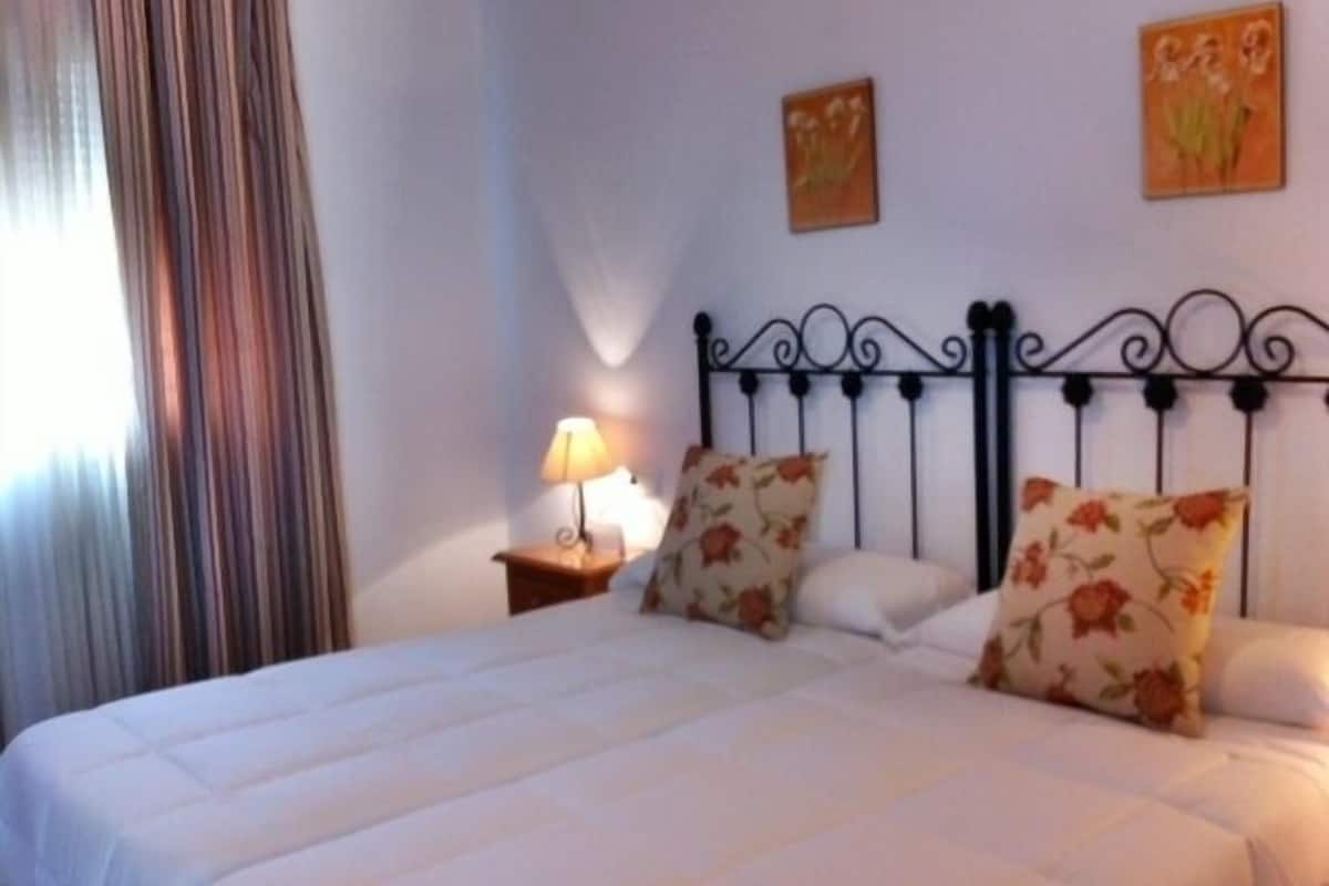 Un lit bien fait, agrémenté de coussins à motifs floraux, une lampe de chevet et deux tableaux accrochés au mur.