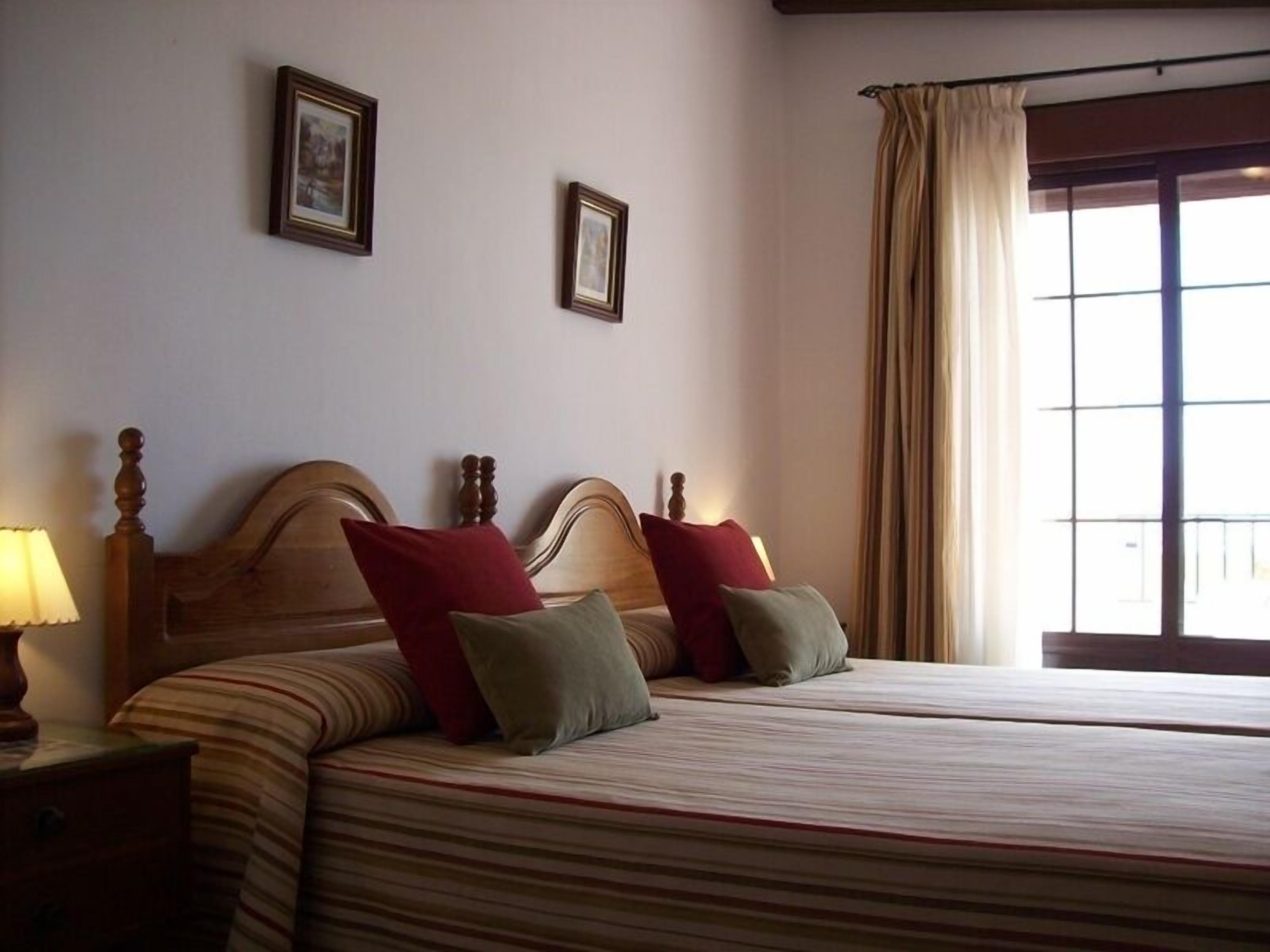 Uma cama de casal com roupa de cama listrada e duas almofadas, uma mesa de cabeceira com abajur e uma janela com cortinas.