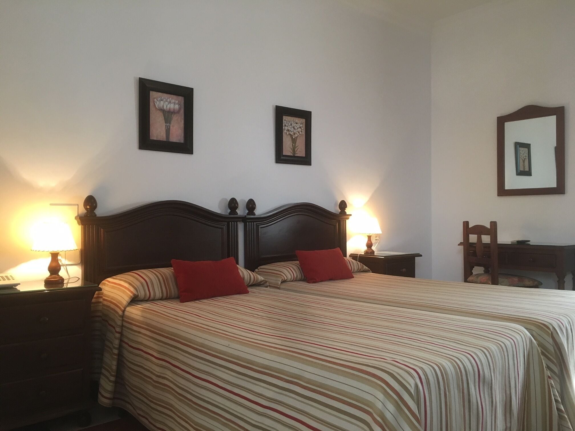 A neatly made bed with striped bedding and two red pillows, flanked by bedside tables with lamps. There are two framed pictures on the wall above the bed.