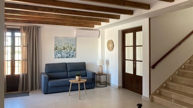 A living room with a blue sofa, a small round table, and a lamp. There is a staircase to the right and a door with glass panels. - Hotel El Molino (Osuna)