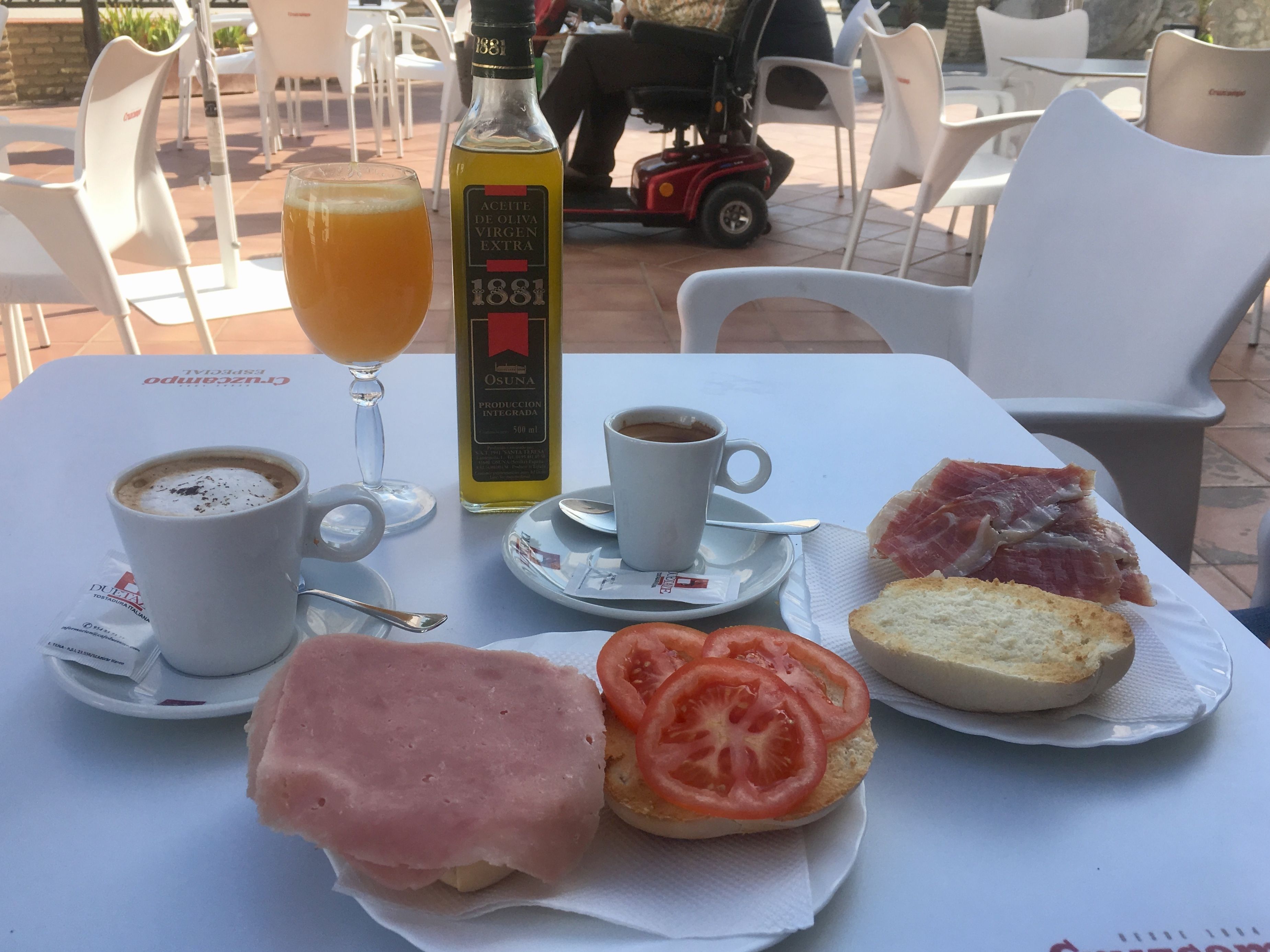 A table with a bottle of olive oil, a glass of orange juice, a cup of coffee, a sandwich with tomato, and a piece of ham.
