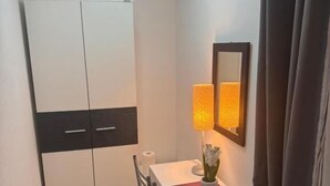 A small, neatly arranged room with a white wardrobe, a desk with a lamp, and a mirror on the wall.