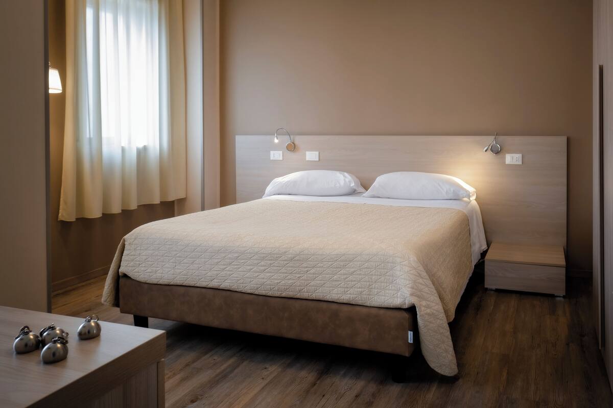 Una cama bien hecha con cubrecama acolchado, una mesita de noche con lámpara y una ventana con cortinas.