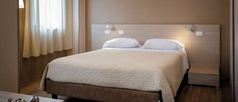 A neatly made bed with a quilted cover, a bedside table with a lamp, and a window with curtains.