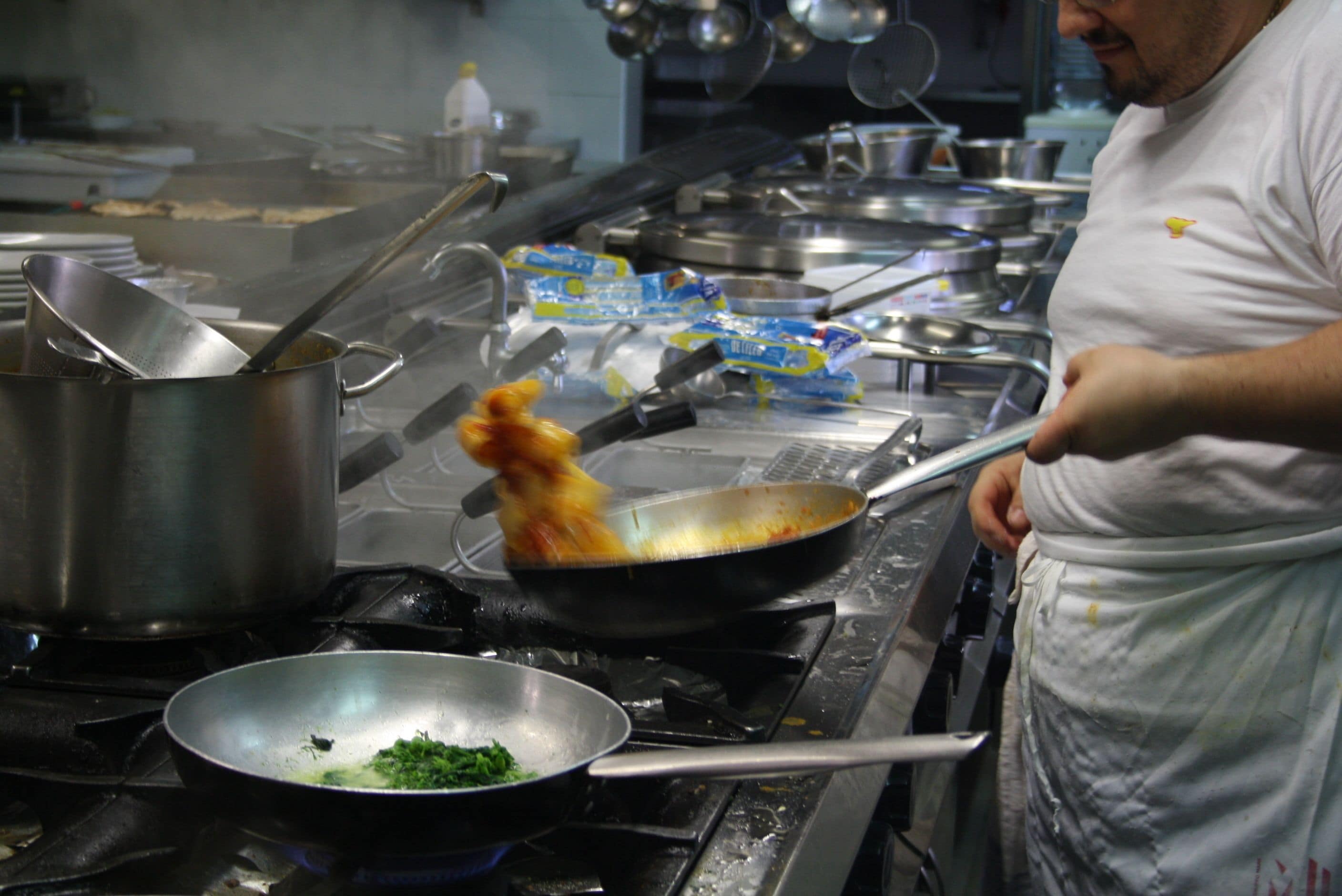 Una cucina con pentole e padelle in acciaio inossidabile, uno chef in divisa bianca e diversi utensili da cucina.