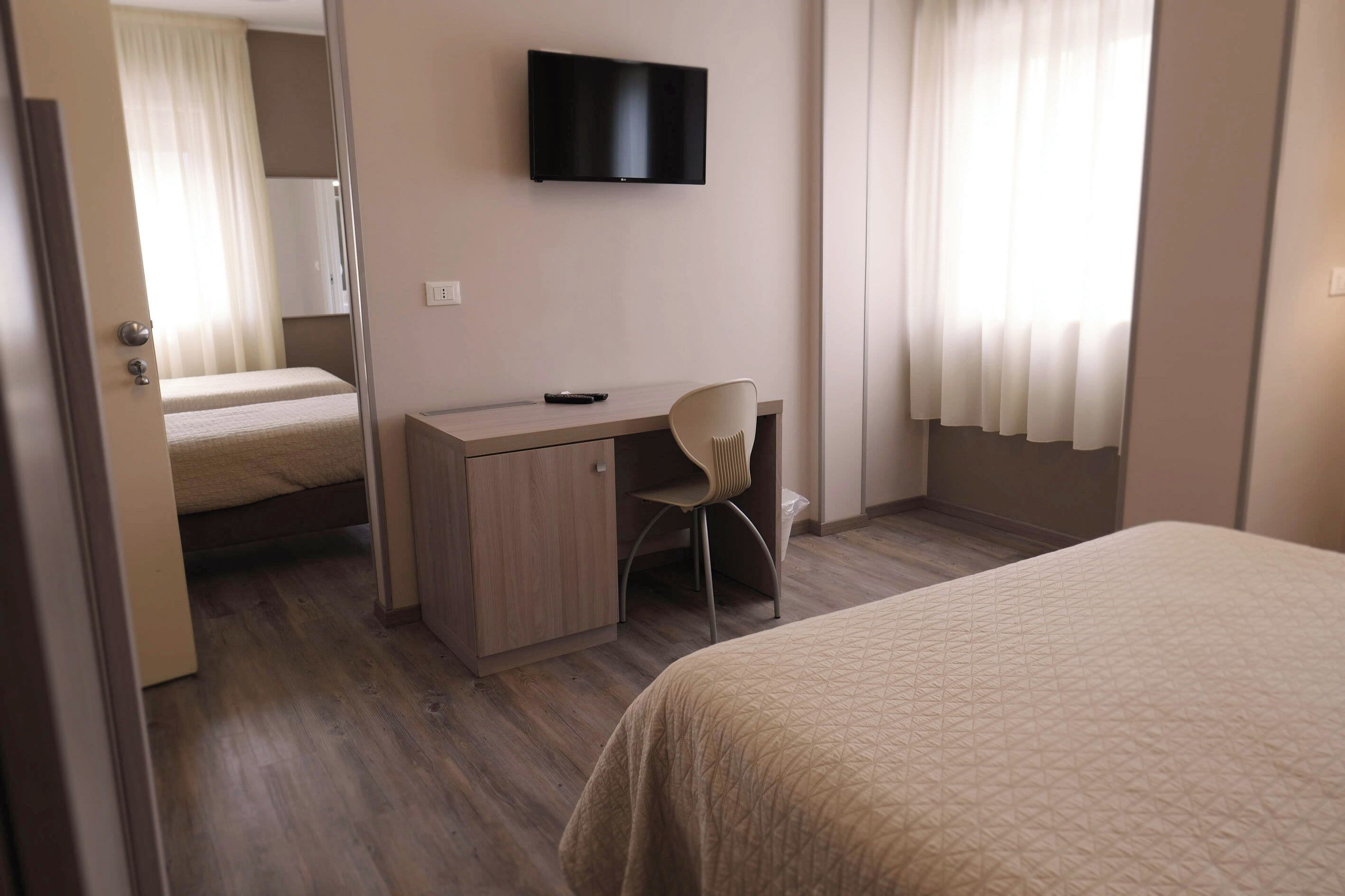 A hotel room with a bed, a desk with a chair, a television on the wall, and a window with curtains.
