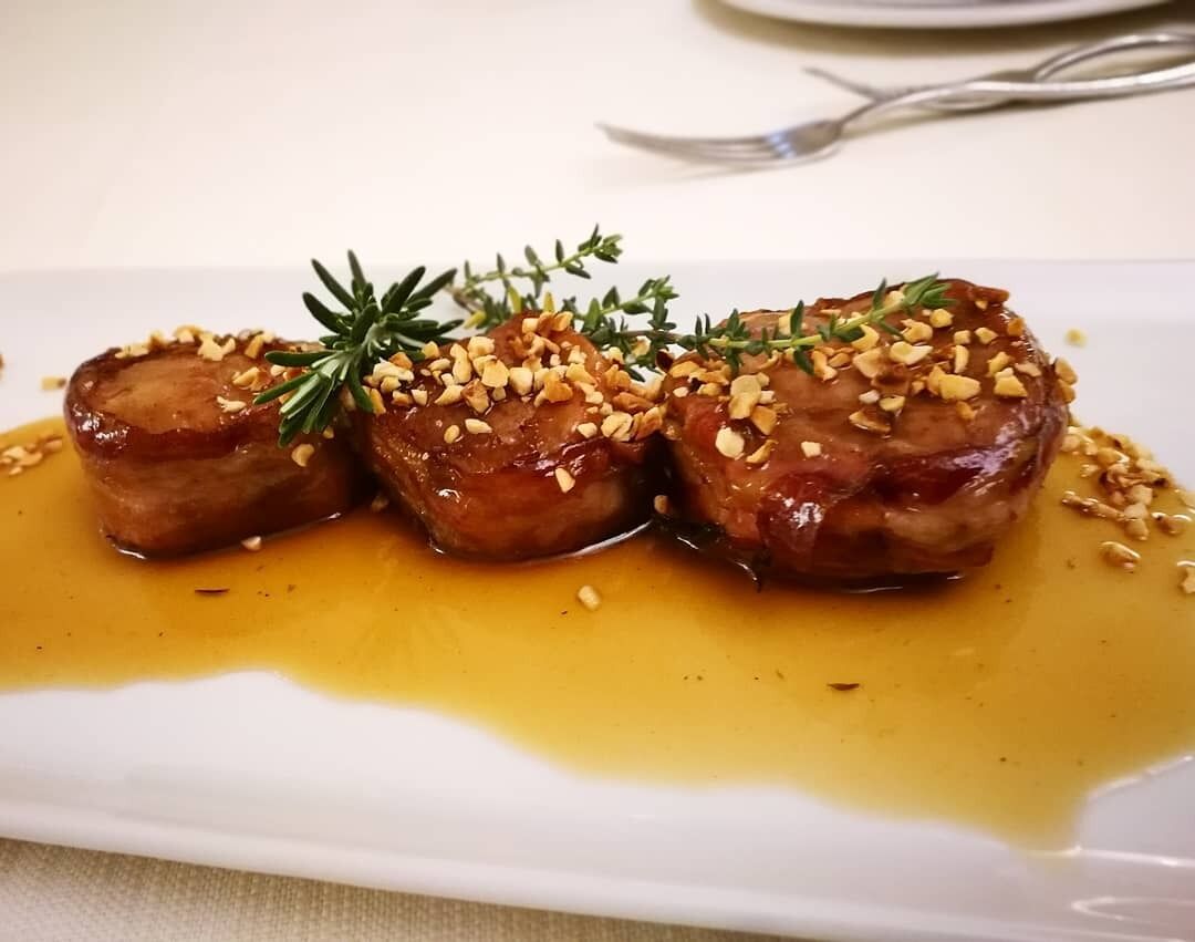 Un piatto con tre fette di carne guarnite con pignoli e un rametto di rosmarino, servito su un piatto bianco con un fondo di salsa.