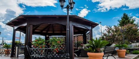 A gazebo with a metal roof, surrounded by chairs and potted plants, under a clear sky.