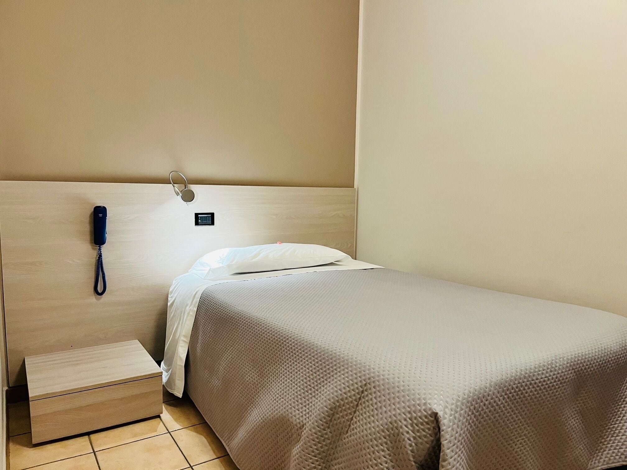 Een klein, netjes opgemaakt bed met een houten hoofdbord, een telefoon op het nachtkastje en een klein nachtkastje.