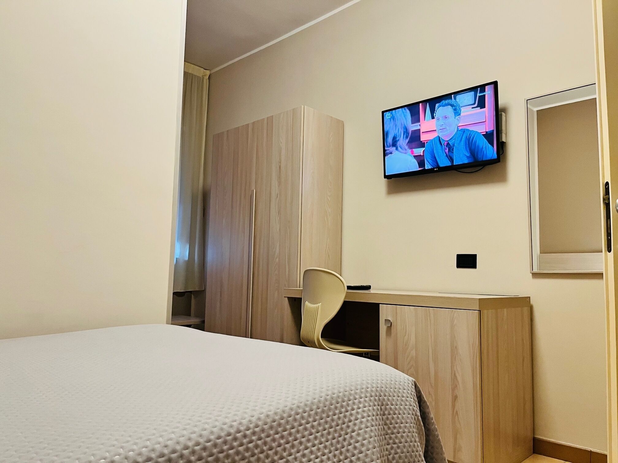 Une chambre d’hôtel équipée d’un lit, d’un bureau, d’une chaise, d’une armoire et d’un téléviseur fixé au mur.