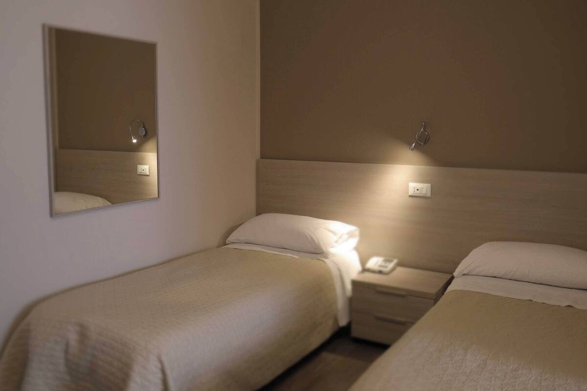 Habitación de hotel con dos camas individuales, una mesita de noche con teléfono, un espejo y una lámpara de pared.