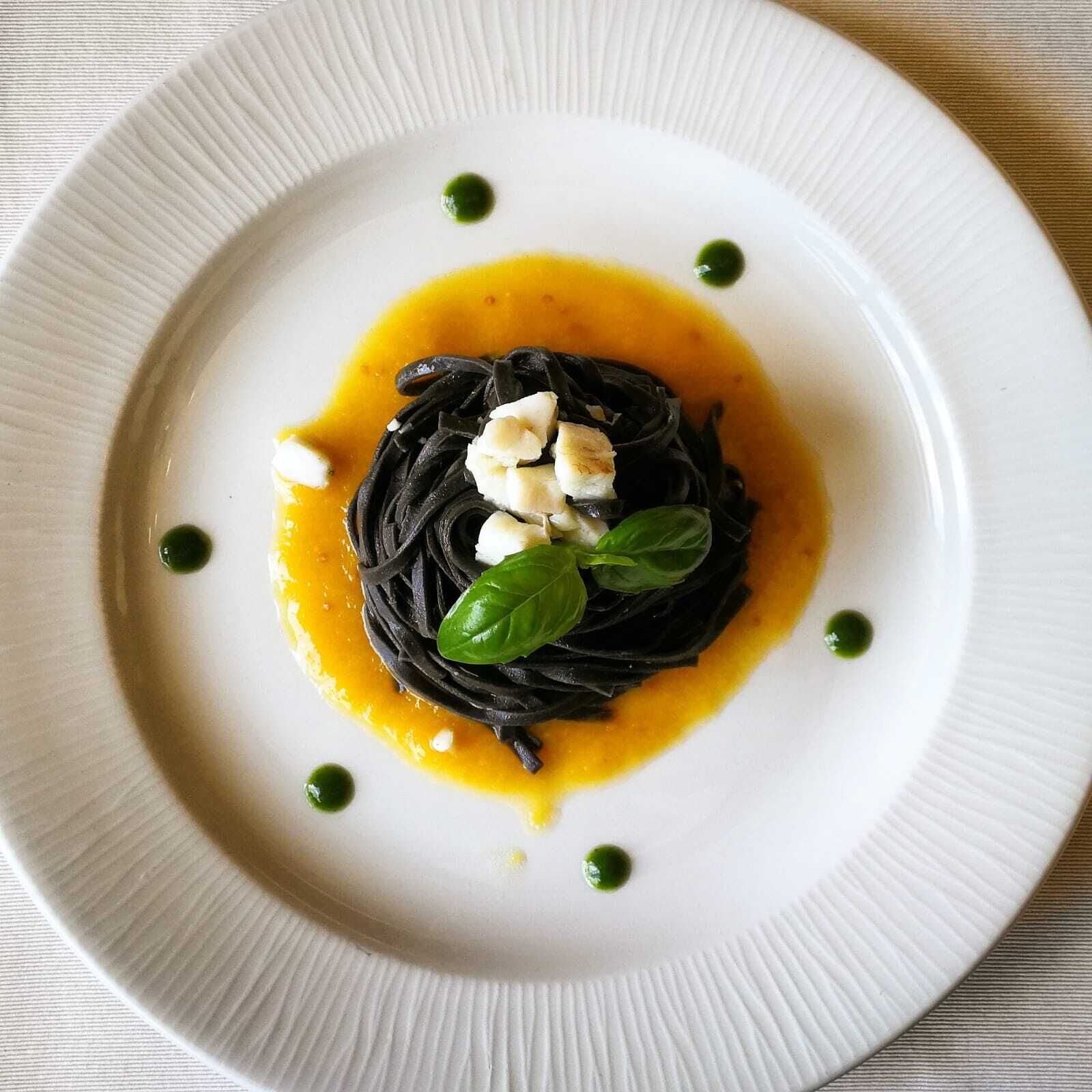 Une assiette de pâtes noires nappées d’une sauce jaune, garnie de basilic et de fromage blanc, le tout présenté sur une assiette blanche.