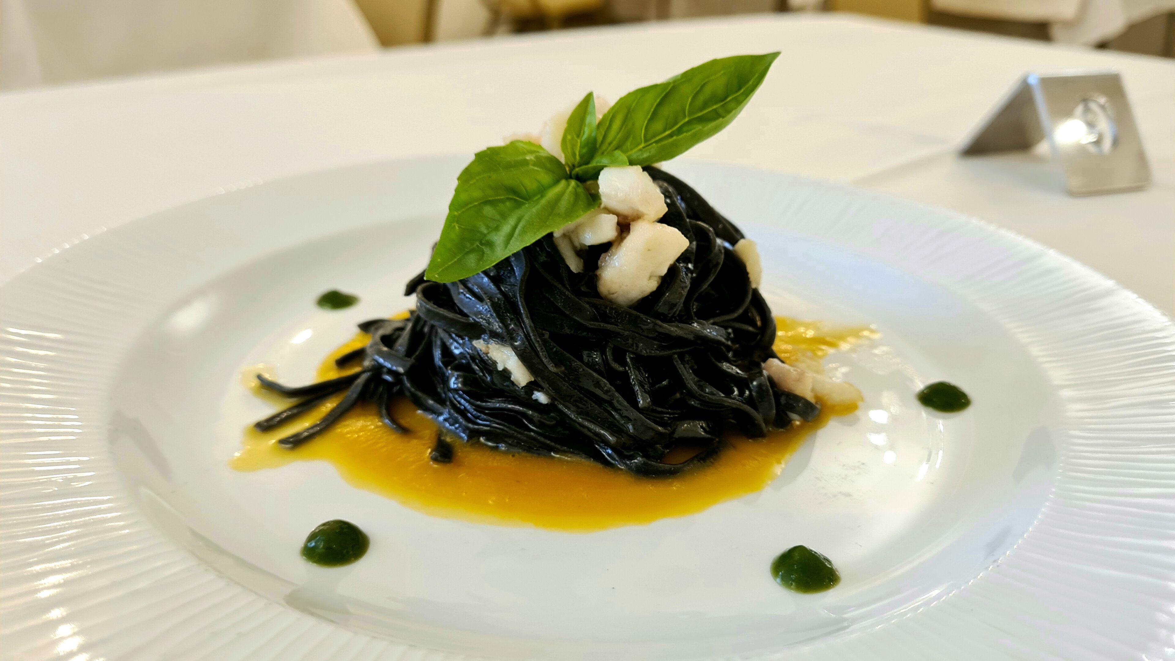 Plato de pasta negra, adornado con albahaca y una cucharada de crema, sobre un plato blanco con salsa amarilla y puntos verdes.