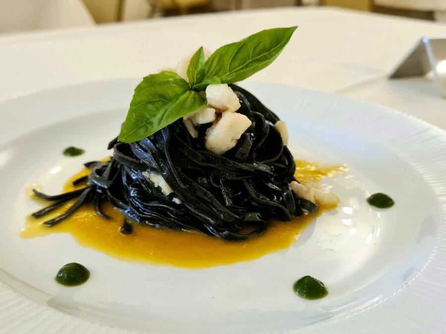 Prato de massa preta com um toque de manjericão e um pouco de creme, servido em prato branco com molho amarelo e pontos verdes.