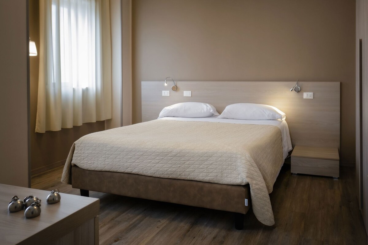 Una cama bien hecha con cubierta acolchada, cabecera de madera, lámparas de noche y mesita de noche con hervidor.