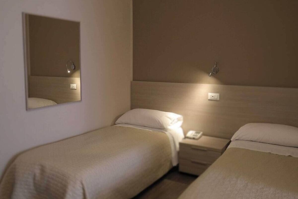 Habitación de hotel con dos camas individuales, una mesita de noche con lámpara, un espejo y un aplique en la pared.