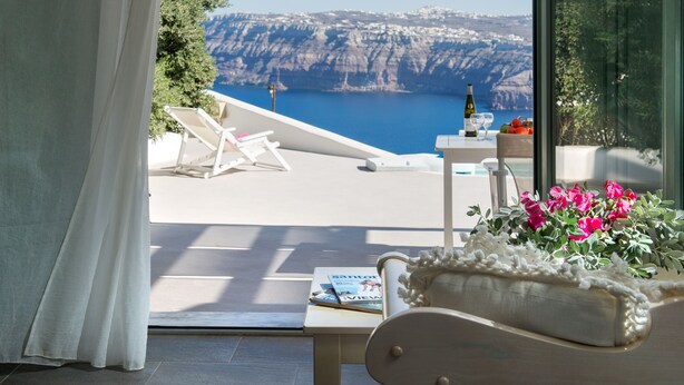 A balcony with a view of a cliff and sea, a table with a bottle and glasses, and a chair with a magazine. - Aura Marina Apartments Santorini (Santorini)