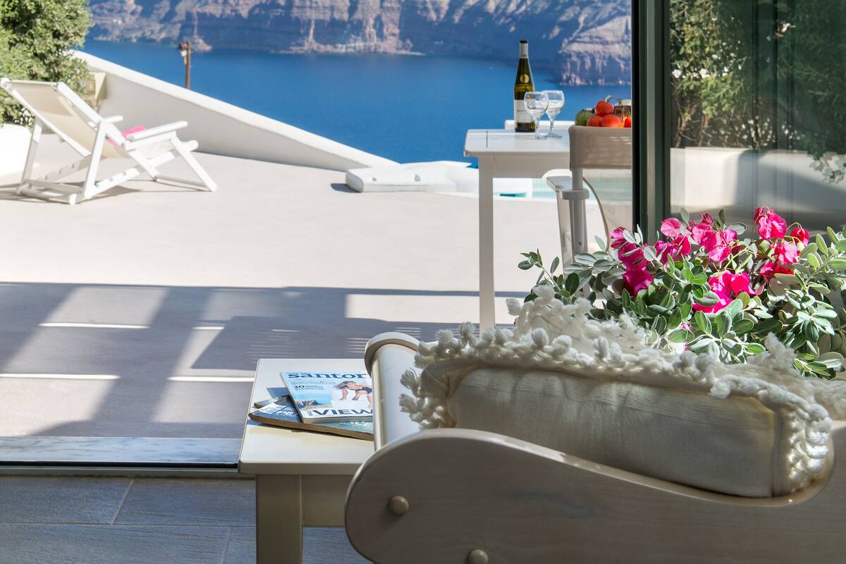 Un balcon donnant sur une falaise et la mer, une bouteille de vin, un verre et une table avec des magazines et des fleurs.