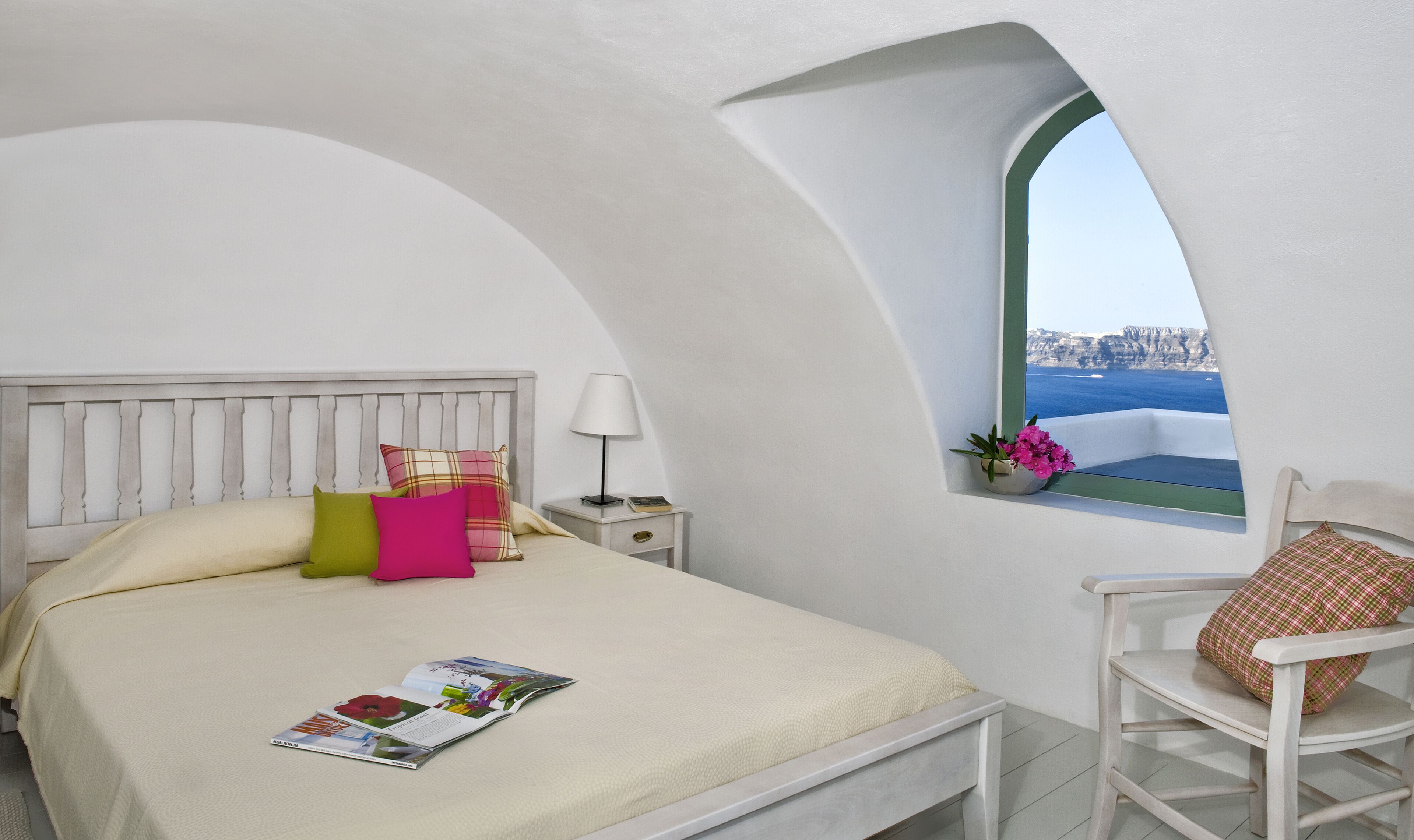A bed with a magazine, a nightstand with a lamp, a chair, and a window with a view of the sea.
