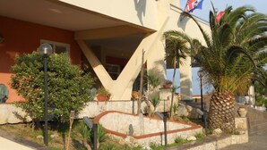 A building with a modern architectural design, featuring a prominent triangular structure and multiple balconies. There are several flags displayed, including the Brazilian flag. The surrounding area includes well-maintained landscaping with various plants and a palm tree. - Le Dune (Castel Volturno)