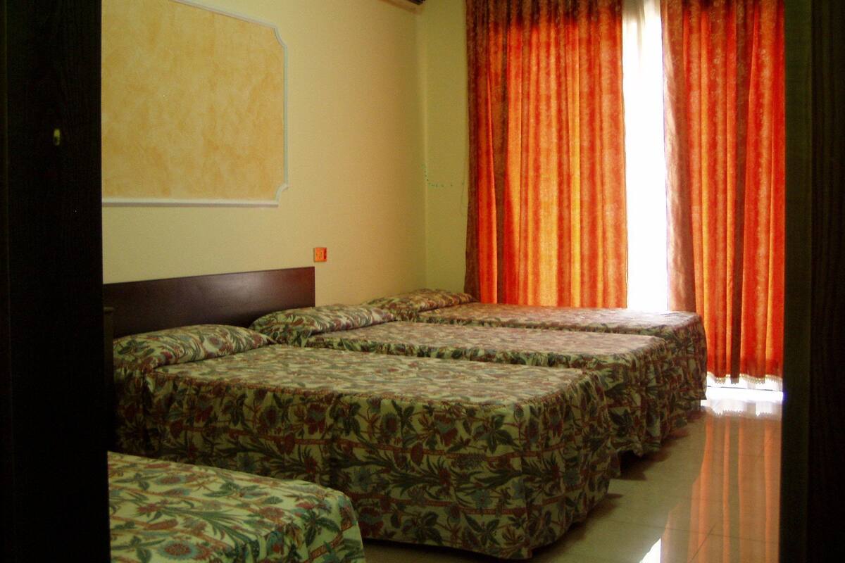 A hotel room with four beds, a large window with orange curtains, an air conditioning unit on the wall, and a plain wall with a small picture.