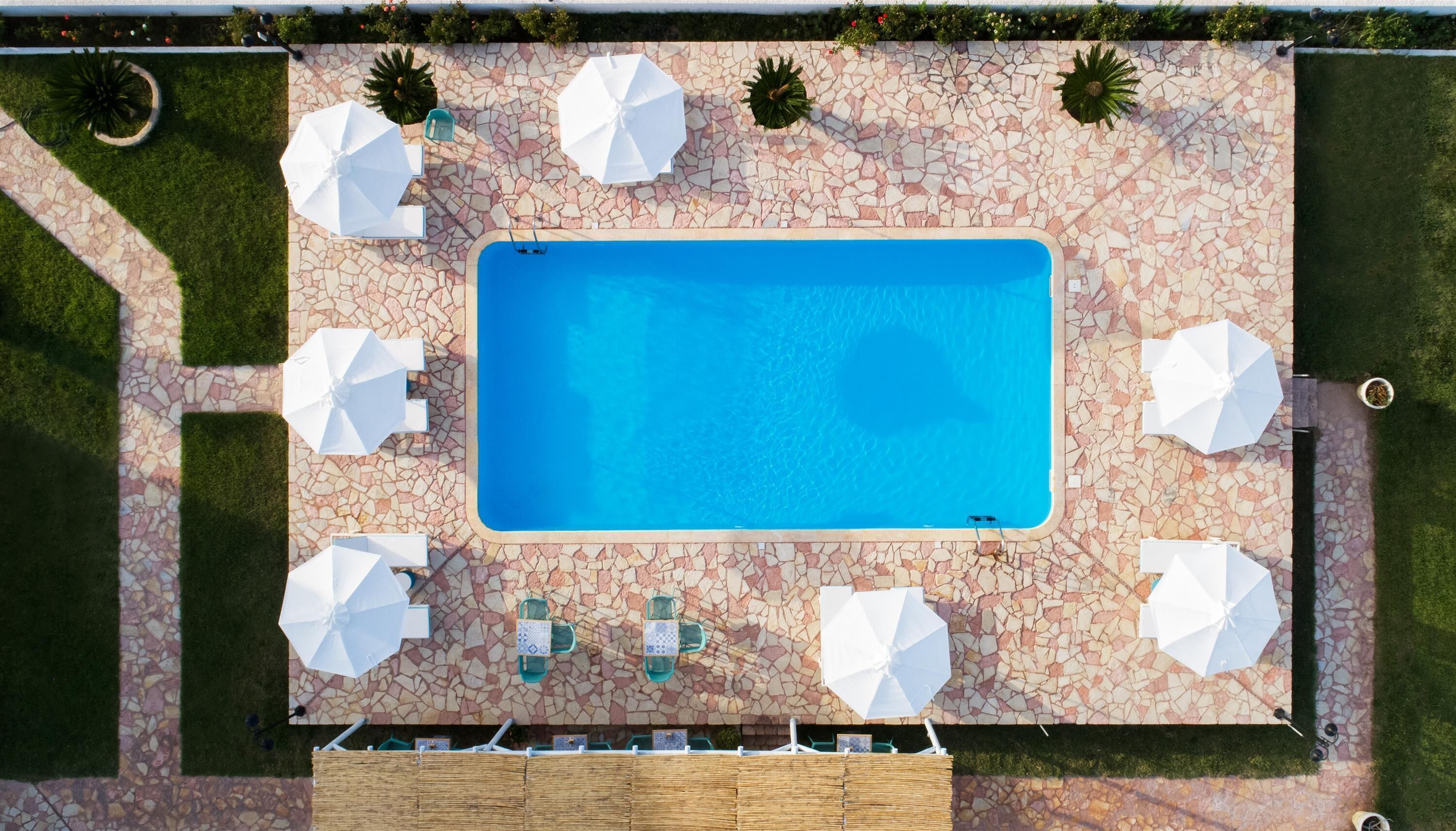 Vue aérienne d’une piscine rectangulaire entourée d’une terrasse dallée de mosaïques, agrémentée de parasols blancs et de chaises.