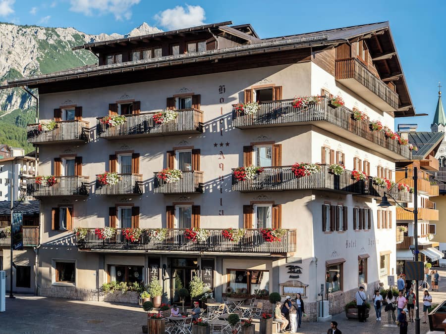 Un hotel de varios pisos con balcones decorados con flores, un paisaje montañoso al fondo y una torre del reloj.