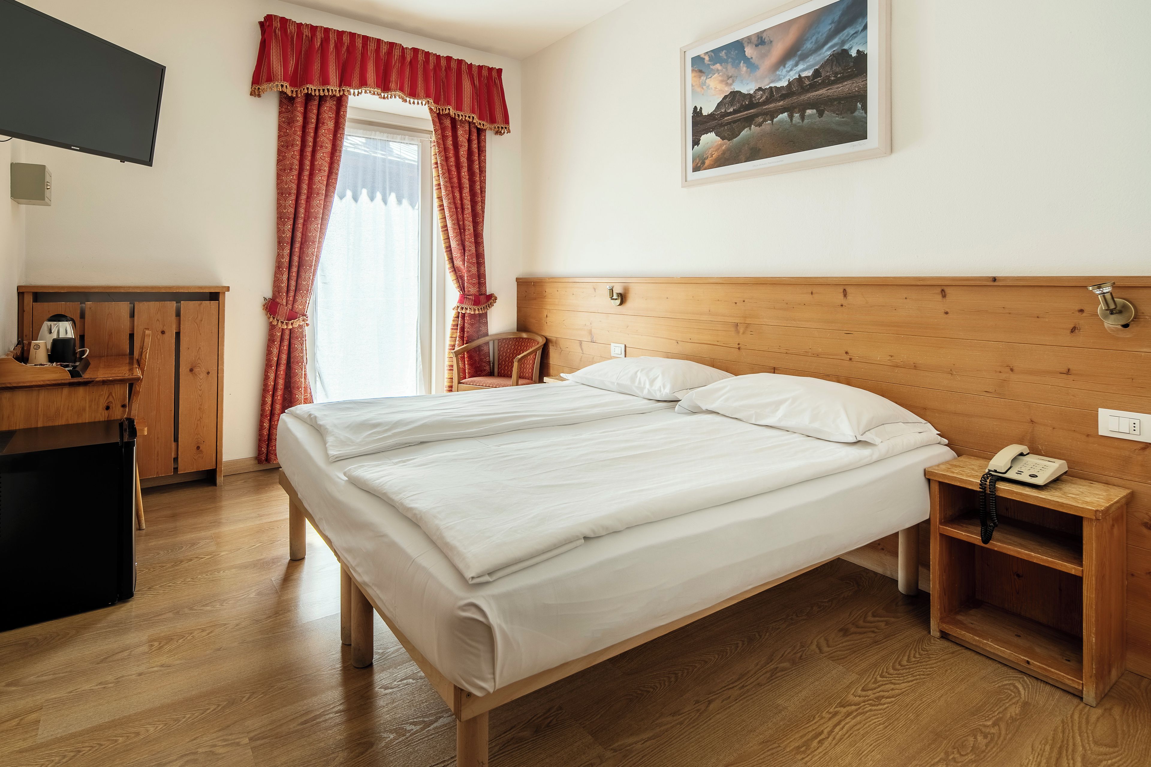 Une chambre d’hôtel avec un lit en bois, un téléphone sur la table de chevet, une commode en bois, une chaise, un bureau avec une bouilloire et un tableau au mur.