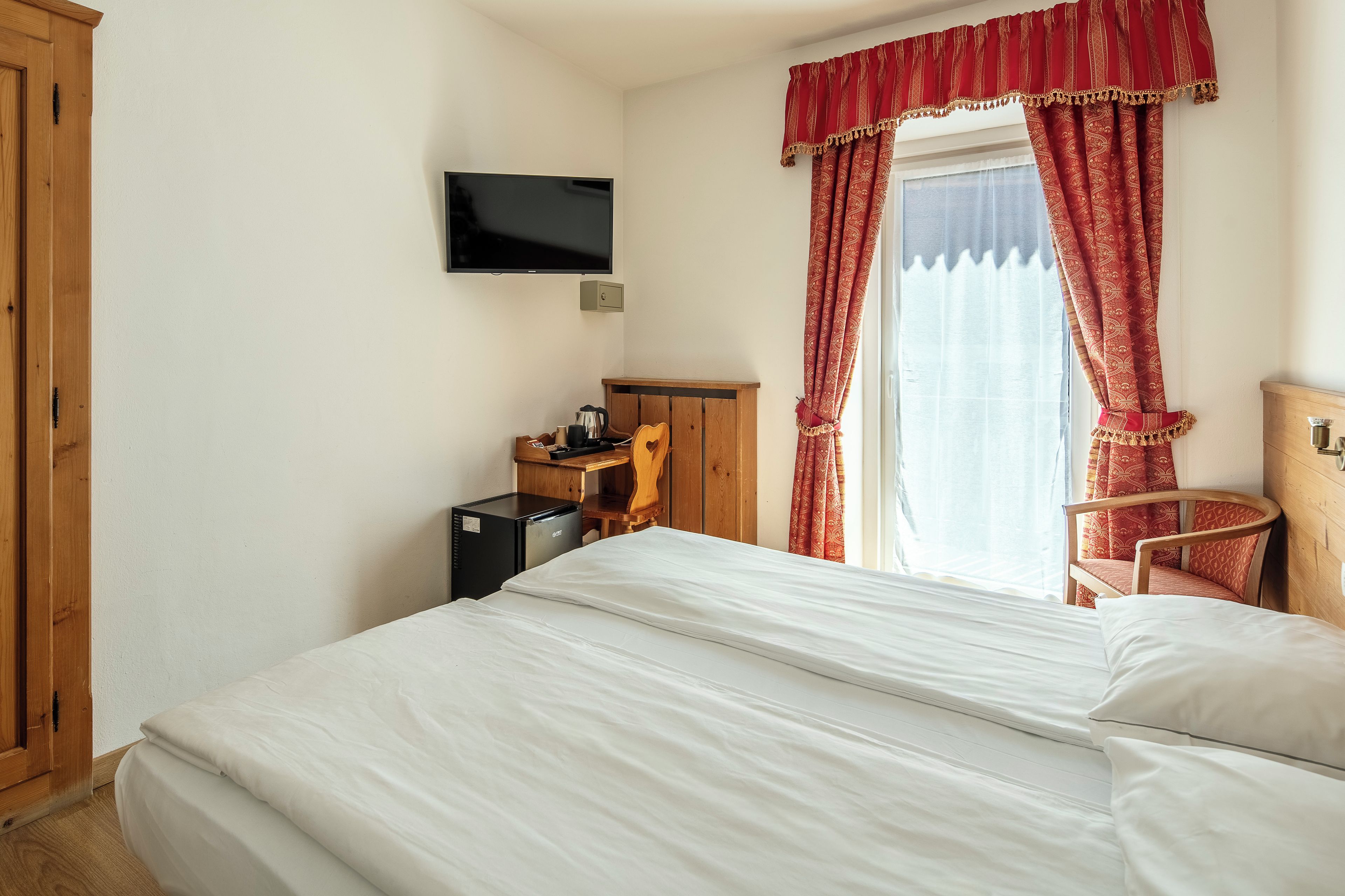 A hotel room with a bed, a television mounted on the wall, a desk with a kettle, a chair, and a window with red curtains.
