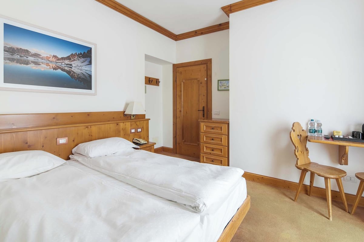 Habitación de hotel con cama, escritorio con silla, armario de madera, puerta de madera y un cuadro en la pared.