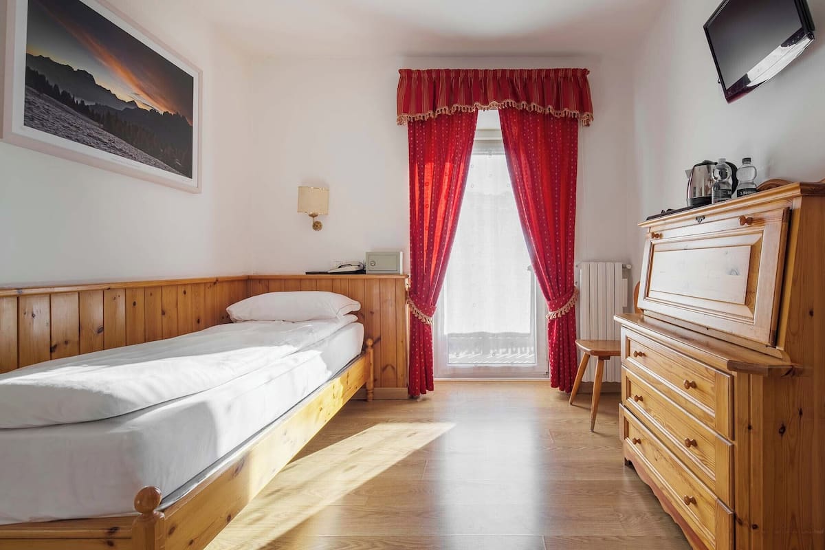 Un dormitorio con cama, cómoda de madera, televisor montado en la pared, ventana con cortinas rojas y un cuadro enmarcado.
