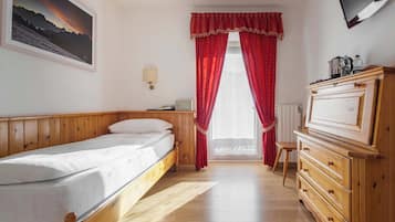 Un dormitorio con cama, cómoda de madera, televisor montado en la pared, ventana con cortinas rojas y un cuadro enmarcado.